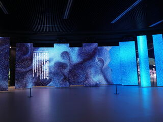 In einem dunklen Raum wird eine Kunstinstallation aus mehreren quadratischen Säulen in unterschiedlichen Größen ausgestellt. Die Säulen bestehen aus LED-Displays und zeigen Bilder in verschiedenen Blautönen.