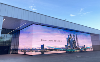 Ein modernes Gebäude mit einem großen digitalen Bildschirm an der Seite, der eine Stadtlandschaft mit Wolkenkratzern und dem Slogan „PIONEERING FOR YOU“ zeigt. Der Himmel ist klar mit ein paar Wolken. Der Bereich vor dem Gebäude ist gepflastert und leer.