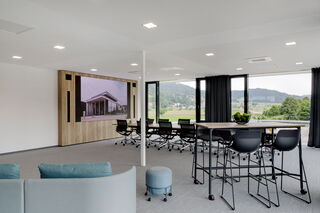 Der neue Prinzbach Anbau Ein modernes helles Bürogebäude von innen ausgestattet mit schwarzen Stühlen, einem Stehtisch und einem großen Konferenztisch beide aus Holz. Direkt neben der weitläufigen Glasfront wurde in einem Holzschrank ein LED-Display verbaut.