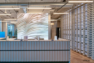 Eines modernen Bürogebäudes, in dem eine LED-Wand direkt im Eingangsbereich hinter einer Empfangstheke installiert wurde. Die Theke aus Steinplatte und wellenförmigem Holzunterbau steht zwischen einer hellen Fensterfront und einer kunstvollen Steinwand.