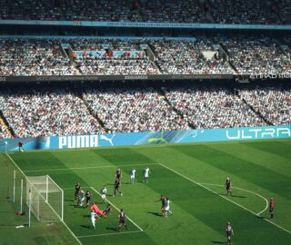 Das Bild zeigt einen Ausschnitt aus einem Fußballspiel aus dem Etihad Stadium in Manchester. Vorne sieht man die beiden Mannschaften auf das Tor schießen. Im Hintergrund sieht man die vollbesetzte Tribüne und eine LED-Doppelbandensystem mit Puma-Werbung.